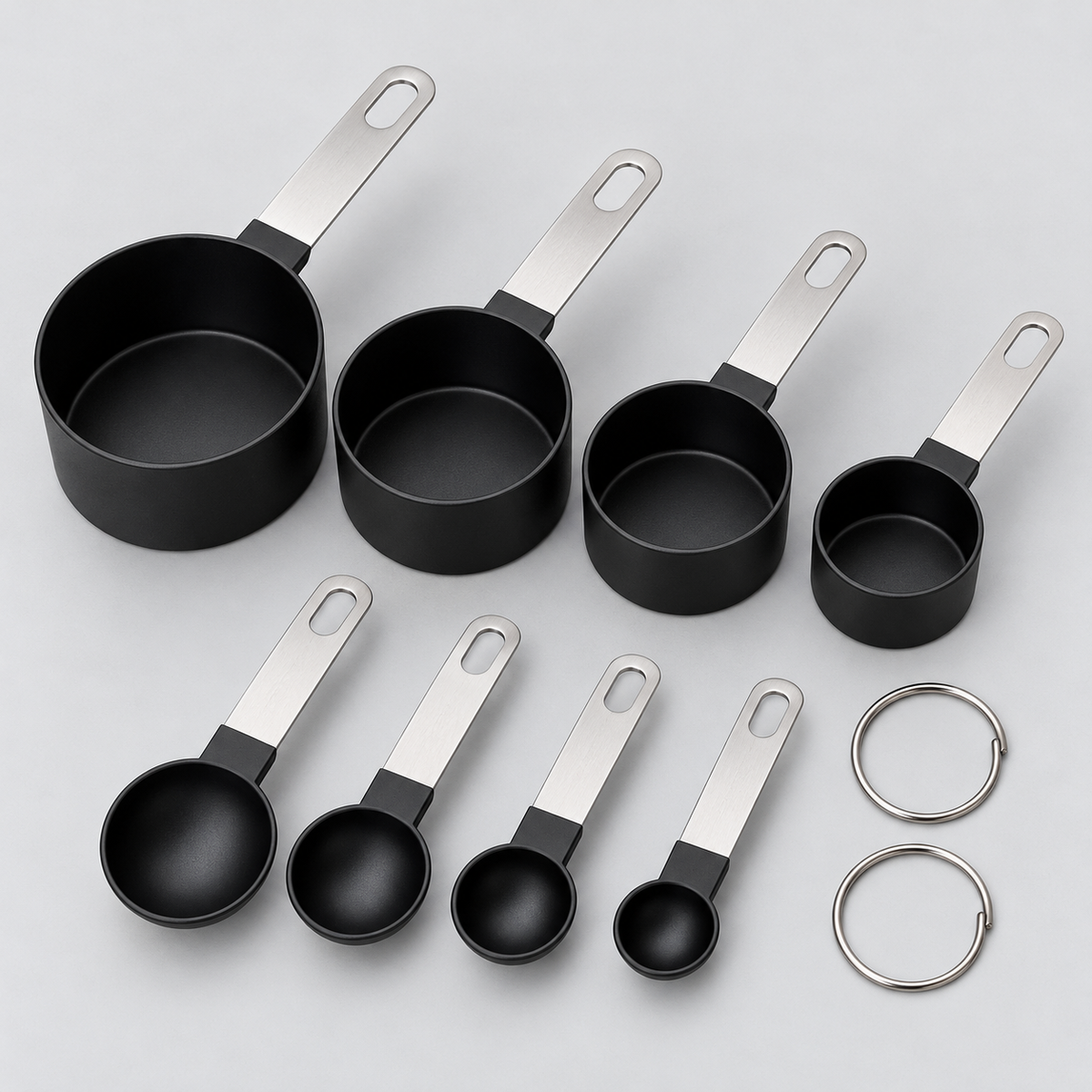 Vista del producto: set negro de cuatro tazas medidoras de plástico cilíndricas con mangos planos plateados de acero y cuatro cucharas medidoras negras redondas con mangos planos plateados, dos anillos metálicos de unión. Ambientación no incluida.